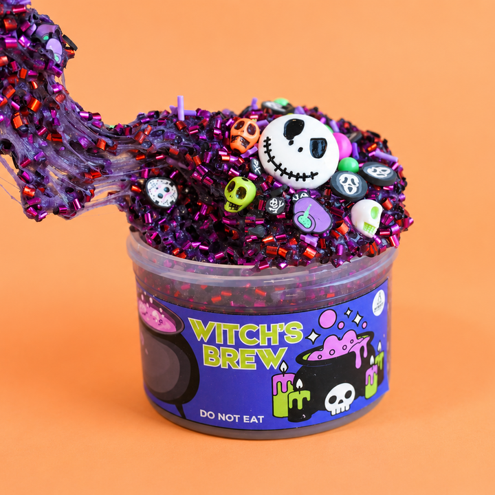 A hand stretches a glossy purple glittery bingsu slime from a clear plastic tub labeled 'Witch's Brew' against a solid orange background. The slime is decorated with colorful Halloween-themed charms, including small purple skulls