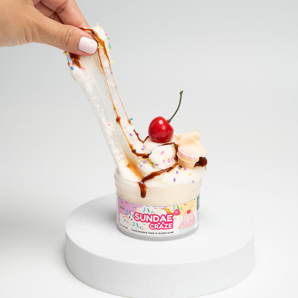 Ice cream sundae with a cherry and chocolate sauce, held by a hand, on a white background.DIY Sundae Craze slime kit with vanilla cloud slime, chocolate slime drizzle, sprinkles, and an ice cream cone charm.