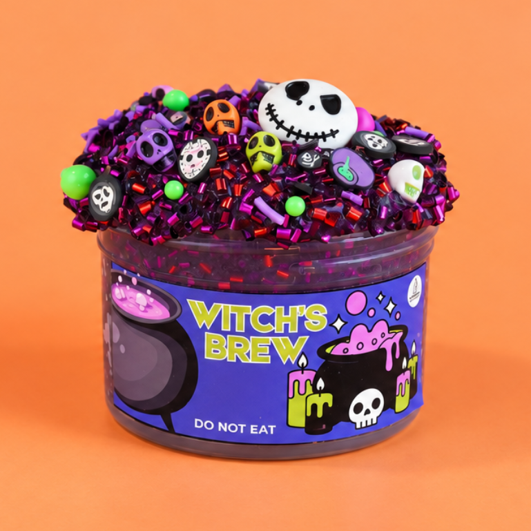 A Halloween-themed slime bucket labeled 'Witch's Brew' on a solid orange background. The slime inside is purple, topped with purple glitter, small colorful skulls, and a white skull topper.